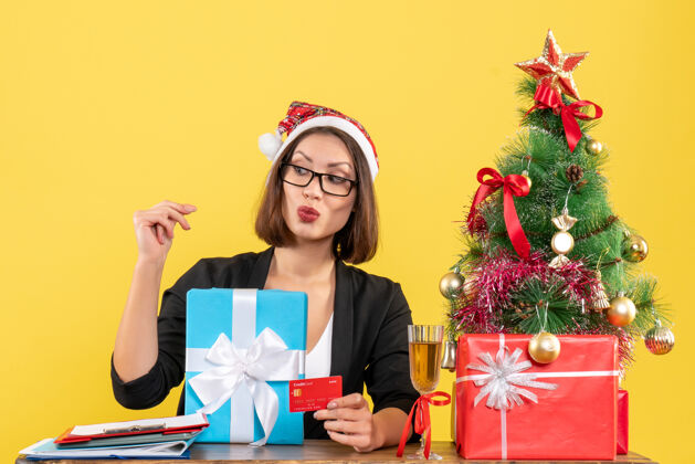 肖像迷人的女士穿着西装 戴着圣诞老人的帽子和眼镜 展示着礼物和银行卡 在黄色的办公室里指着后面指向黄色时尚