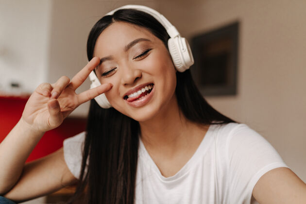 科技可爱的亚洲女人在白色上衣和耳机显示舌头 和平标志和闭上眼睛的姿势女人房子电话