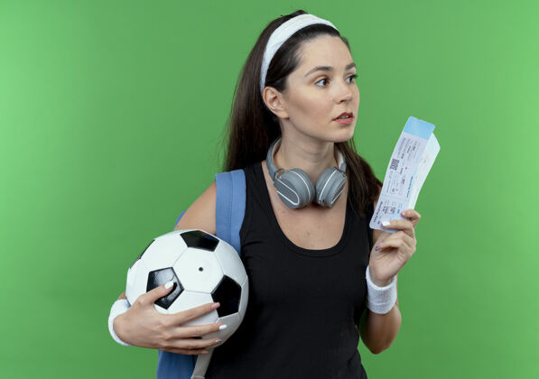 困惑戴着头带 背着耳机 背着背包 拿着足球和机票 困惑地站在绿色背景下 一边看一边的年轻健身女士背包持有足球
