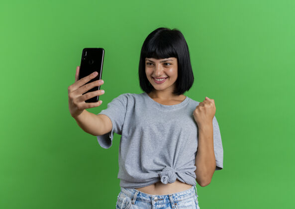 复制年轻的黑发白人女孩微笑着握紧拳头 看着绿色背景上的手机 手机上留有复印空间女孩空间抱着