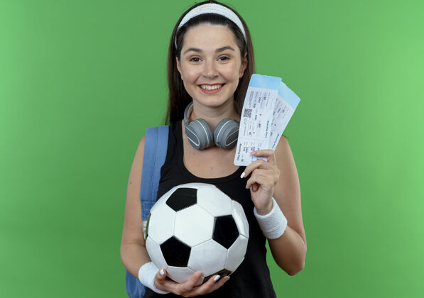 立场戴着头戴式耳机 背着背包 拿着足球和机票的年轻健身女士站在绿色背景下 面带微笑地看着摄像机举行票相机