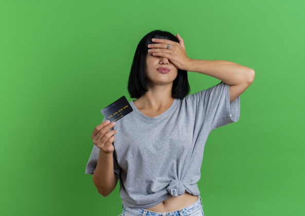 手一个年轻的黑发白人女孩拿着信用卡 闭上眼睛 手被隔离在绿色背景上 留着复印空间持有深色不愉快