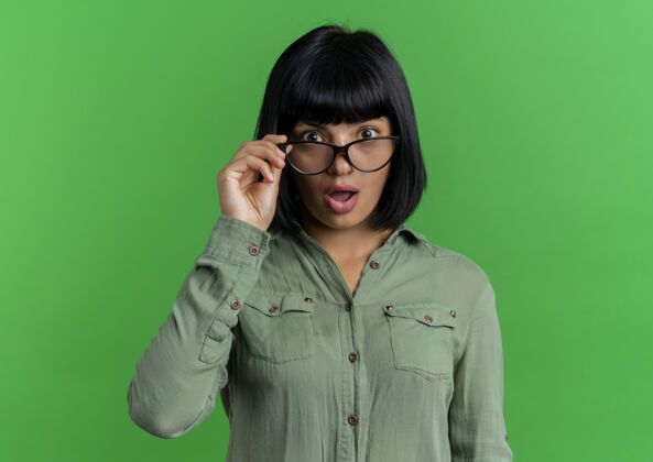 复制震惊的年轻黑发白人女孩手持光学眼镜 看着隔离在绿色背景和复印空间的相机女孩年轻黑发