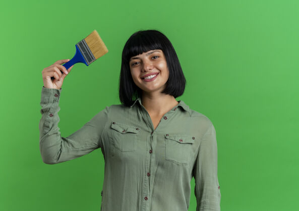年轻微笑着的年轻黑发白人女孩拿着画笔在绿色背景上与复制空间隔离油漆深色复制