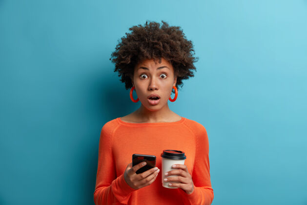 短信惊呆了的少数民族妇女在网上看惊艳的新闻 手里拿着手机 收到了促销码 喝着外卖咖啡 穿着橙色的套头衫 对着蓝色的墙壁摆姿势兴奋女人无线