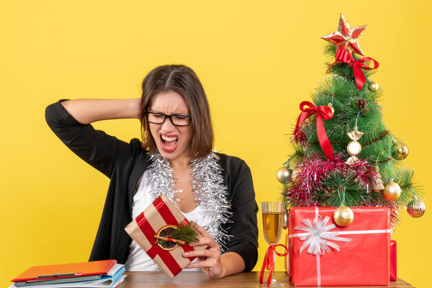 抱着一位穿着西装 戴着眼镜 情绪激动地拿着礼物的美丽商务女士坐在办公室的一张桌子旁 桌上放着一棵圣诞树情绪礼物人