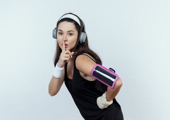 站立戴着耳机和智能手机袖标的戴着头带的年轻健身女士在白色背景上做着手指放在嘴唇上的沉默手势健身臂带制造