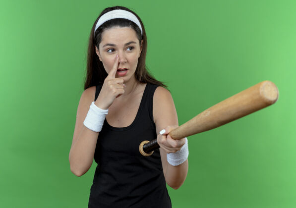 头带戴着头巾的年轻健身女士拿着棒球棒 一边看一边用手指着鼻子站在绿色背景上拿着年轻人健身