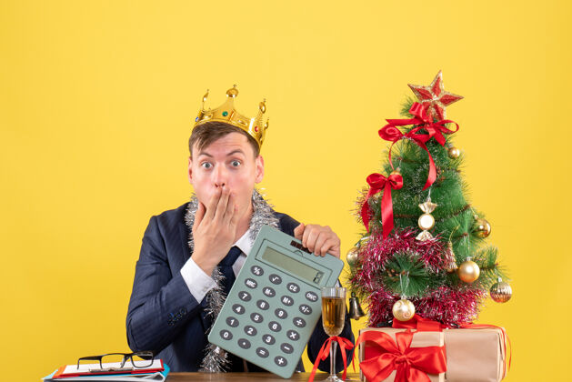 前前视图惊奇的人拿着计算器坐在圣诞树旁边的桌子上 呈现在黄色背景上桌子人树