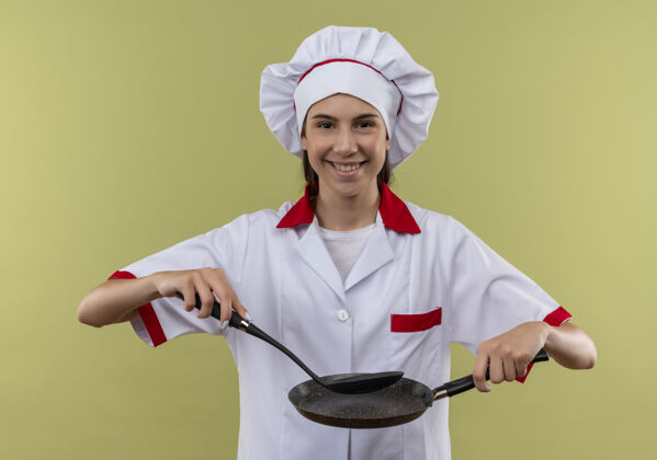 平底锅身着厨师制服 面带微笑的年轻白人厨师女孩手拿煎锅和抹刀 将它们隔离在绿色背景上 并留有复印空间厨师拿着女孩