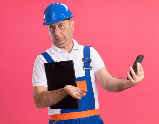 人困惑的成年建筑工人制服举行剪贴板和电话隔离在粉红色的墙上脸电话成人