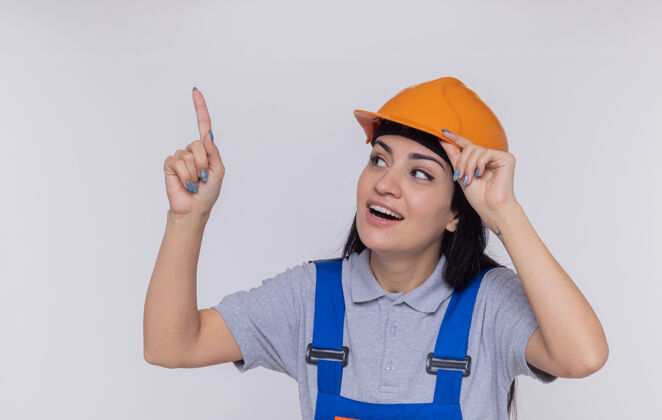 什么身穿建筑制服 头戴安全帽的年轻建筑工人抬起头微笑着用食指指着什么东西建设者微笑向上