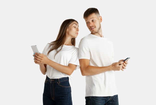 惊奇美丽的年轻夫妇的肖像被隔离在白色的工作室背景上面部表情 人类情感 广告理念都拿着智能手机 女人在屏幕上看着保守秘密人情侣男性