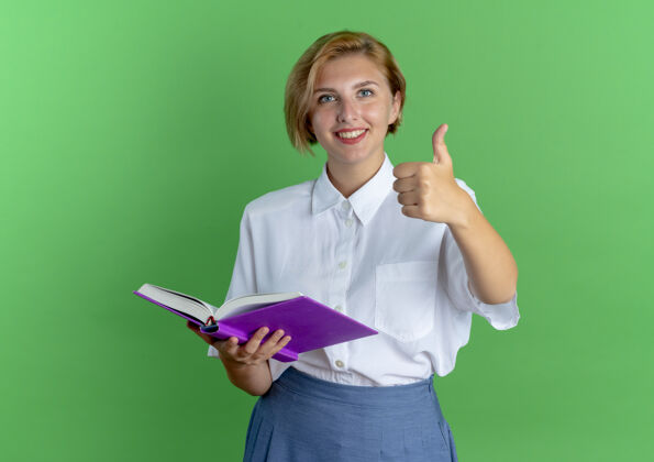 女孩年轻的微笑金发的俄罗斯女孩竖起大拇指拿着一本书在绿色背景上与复制空间隔离年轻金发微笑