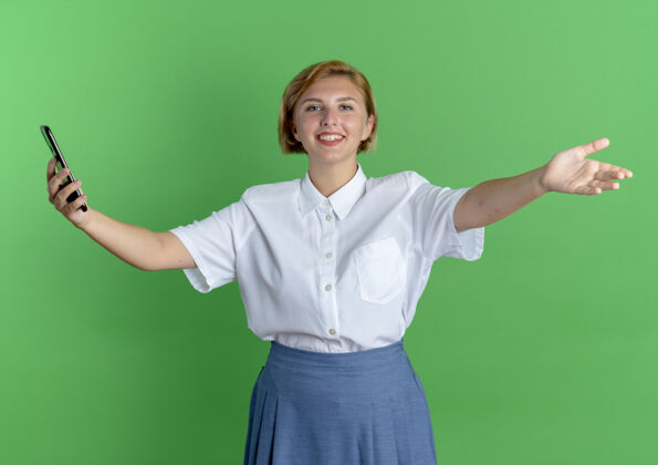 女孩年轻的微笑的金发俄罗斯女孩手持电话 张开双臂 在绿色背景上与复制空间隔离微笑金发手臂
