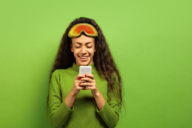 黑发以绿色工作室为背景 黑色皮肤的非洲裔年轻女子的滑雪面具肖像人类情感 面部表情 销售 广告 冬季运动和假期的概念使用智能手机微笑惊喜肖像