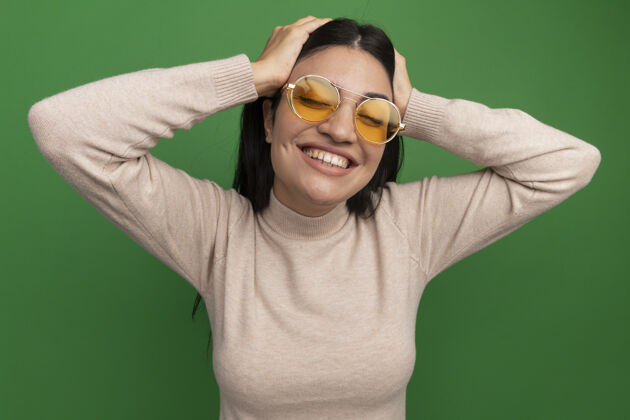 人戴着太阳眼镜的黑发美女微笑着把双手放在隔离在绿色墙上的头上漂亮姿势黑发