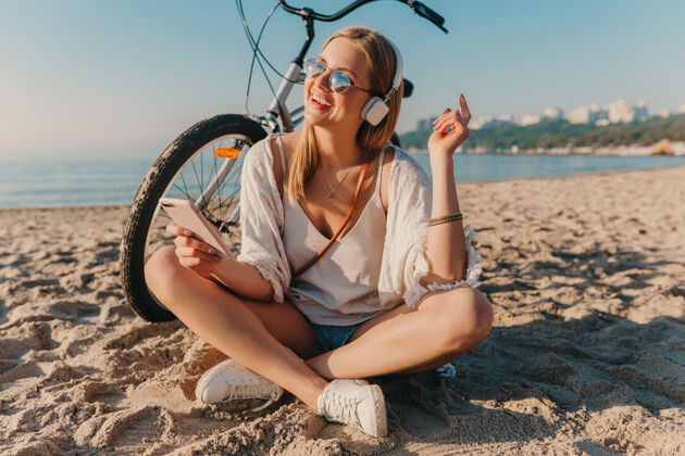 配件时尚年轻迷人的金发微笑的女人坐在沙滩上骑着自行车戴着耳机听音乐美丽休闲海洋