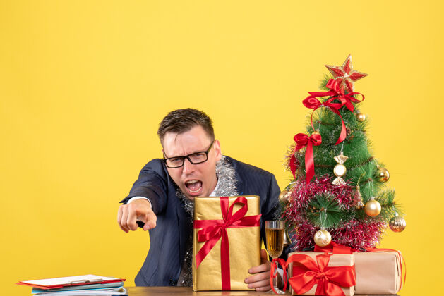 男人前视图愤怒的男子指着摄像机坐在圣诞树旁的桌子上 呈现黄色背景照相机男人礼物