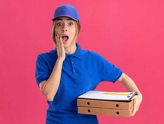 剪贴板穿着制服的年轻漂亮的送货员惊讶地把手放在脸上 把比萨饼盒孤立地放在粉红色的墙上女孩惊喜手