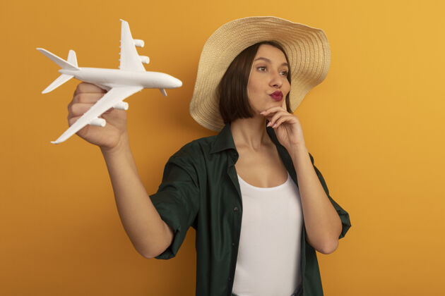 帽子戴着沙滩帽的自信美女把手放在脸上 举着模型飞机看着橙色墙上孤立的一面漂亮脸飞机