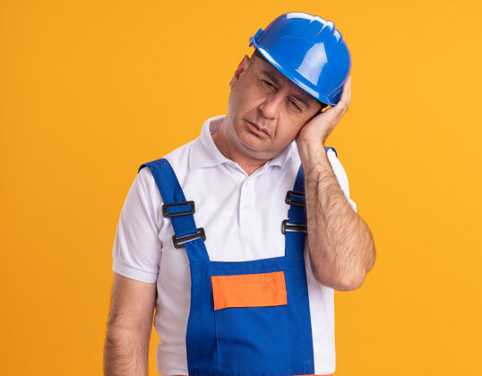 姿势疼痛的成年建筑工人穿着制服把手放在隔离在橙色墙上的头上人表情立场