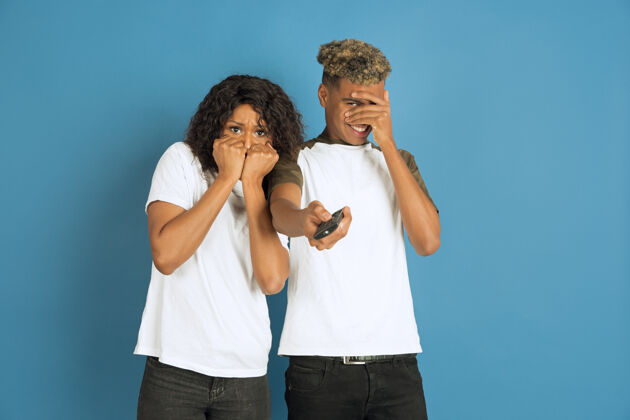 关系年轻感性的非裔美国人 穿着白色衣服 在蓝色背景上摆姿势美丽的情侣人类情感的概念 面部表情 关系 广告一起看电视 看起来很害怕非洲朋友人