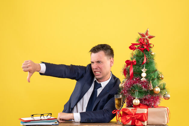 礼物前视图愤怒的年轻人做拇指向下的标志坐在圣诞树附近的桌子和黄色背景上的礼物人人前面