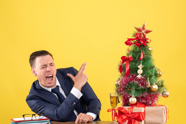 枪前视图愤怒的男子正在制作手指枪标志坐在圣诞树和黄色背景上的礼物附近的桌子上使人礼物