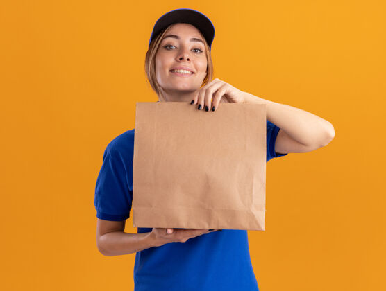 表情微笑着的年轻漂亮的女送货员穿着制服 手里拿着隔离在橙色墙上的纸包女孩人微笑