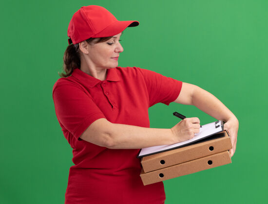 递送身穿红色制服 头戴鸭舌帽的中年女送货员 手里拿着披萨盒和空白页的剪贴板 严肃的脸站在绿色的墙上披萨脸严肃