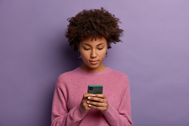 打字外表严肃的非裔美国妇女在现代手机上阅读信息 浏览社交媒体 在显示器上有集中的目光 穿着休闲套头衫非洲裔皮肤网络