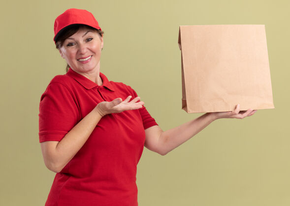 送货身穿红色制服 头戴帽子的中年送货妇女手拿纸包 面带微笑 自信地站在绿色的墙边中间站着帽子