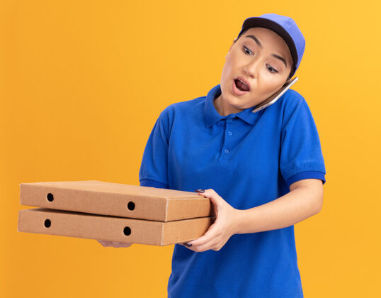年轻人身穿蓝色制服 戴着帽子 拿着披萨盒的年轻送货员站在橙色的墙上 惊讶地用手机交谈帽子送货惊喜