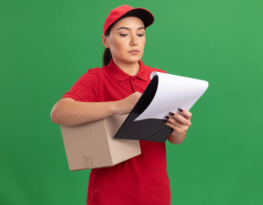 制服年轻的送货员身穿红色制服 戴着帽子 手里拿着纸板箱和空白页的剪贴板 严肃地站在绿色的墙上看着它脸剪贴板帽子