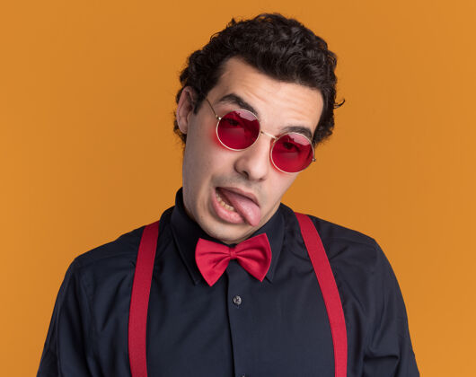 使戴着蝴蝶结 戴着眼镜 系着吊带 站在橘色的墙上做鬼脸 伸出舌头的时髦男人蝴蝶结眼镜吊带