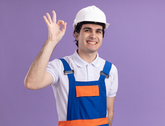 年轻年轻的建筑工人身穿建筑制服 戴着安全帽 面带微笑 站在紫色的墙上 脸上挂着“ok”的牌子头盔表演好