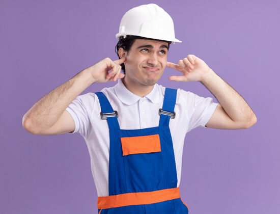 站身穿施工制服 头戴安全帽的年轻建筑工人站在紫色的墙上 用手指捂住耳朵 脸上露出恼怒的表情男人建筑封闭