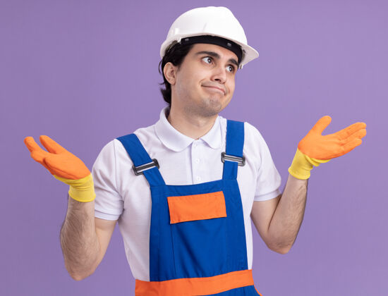 橡胶年轻的建筑工人穿着建筑制服 戴着橡胶手套戴着安全帽 站在紫色的墙上 困惑而不确定地看着前方 举起双臂 没有回答站立头盔手套