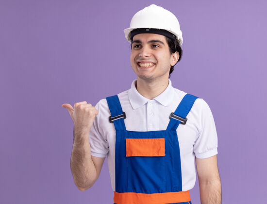 年轻人年轻的建筑工人穿着建筑制服 戴着安全帽 面带微笑地看着一边站在紫色的墙上建筑工人头盔男人
