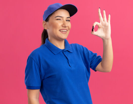 女人身穿蓝色制服 头戴帽子的年轻女送货员站在粉红色的墙上 面带微笑 脸上挂着“ok”的牌子交货站着微笑