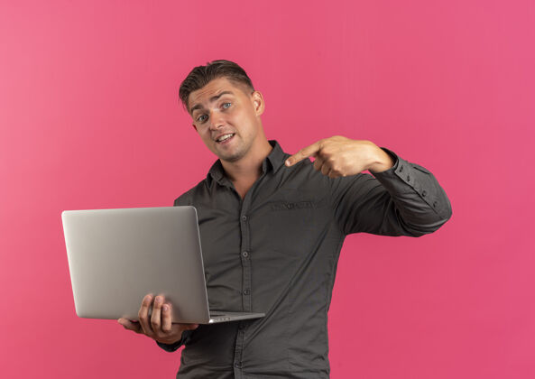 年轻年轻而惊讶的金发帅哥拿着笔记本电脑 指着它 笔记本电脑的背景是粉色的 还有复印空间拿着笔记本电脑帅哥