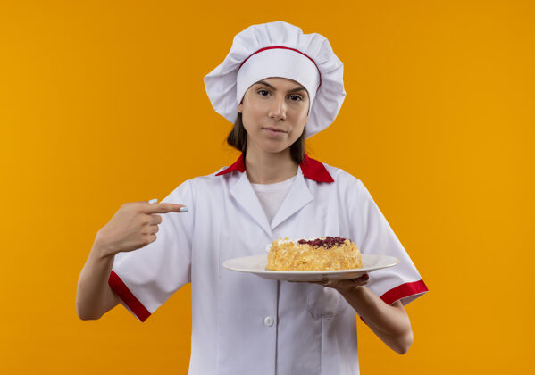 厨师身着厨师制服的年轻自信的白人厨师女孩拿着并指着盘子上的蛋糕 蛋糕被隔离在橙色的背景上 留有复制空间盘子女孩点