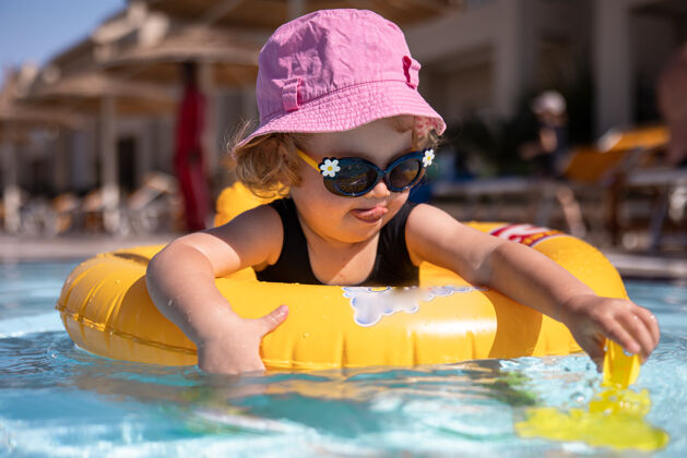 小可爱的小女孩戴着帽子和太阳镜坐在游泳池里玩阳光女孩游戏