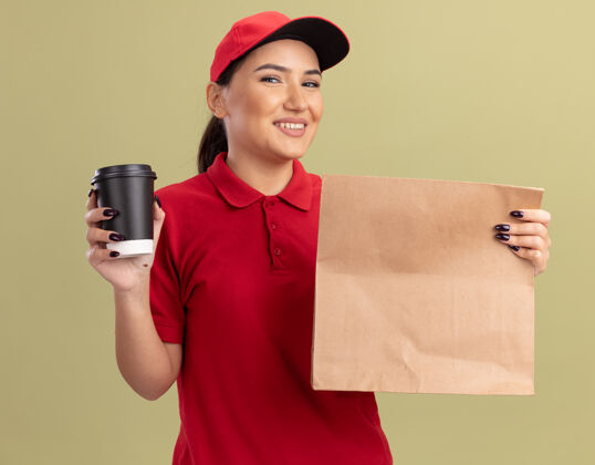 快乐身着红色制服 头戴鸭舌帽 拿着纸包和咖啡杯 脸上挂着微笑 站在绿色墙壁上的快乐年轻女送货员制服微笑帽子