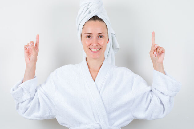 新鲜年轻的女士指着白色浴衣 毛巾和看起来愉快 前视图漂亮女士皮肤