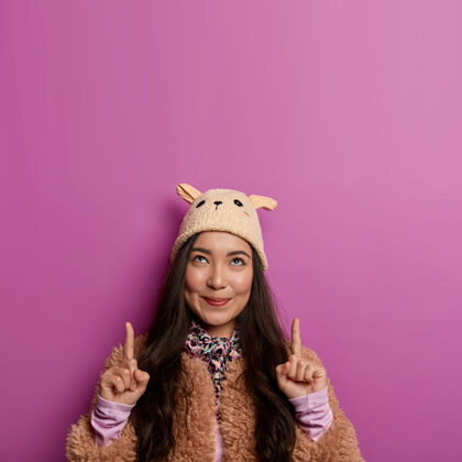 女士可爱女孩的照片指向上面的食指 广告复制空间 显示向上的东西 戴着带耳朵的帽子和棕色外套指标向上韩国人