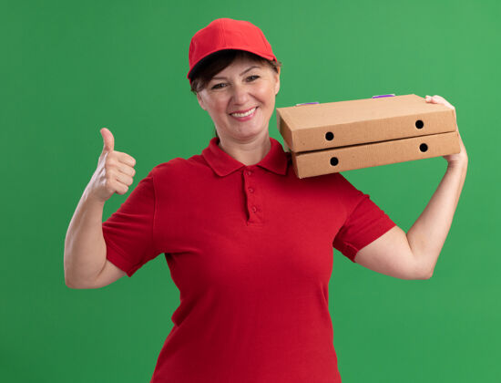 递送快乐的中年送货员 身穿红色制服 戴着帽子 拿着披萨盒 面带微笑 站在绿色的墙上竖起大拇指表演披萨制服
