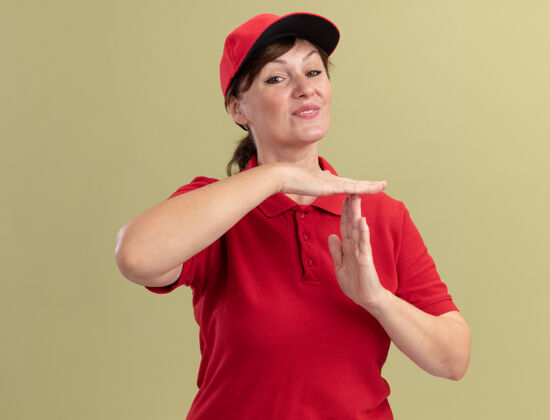 手身穿红色制服 头戴鸭舌帽的中年女送货员微笑着站在绿色墙壁上做着暂停的手势手势中间时间