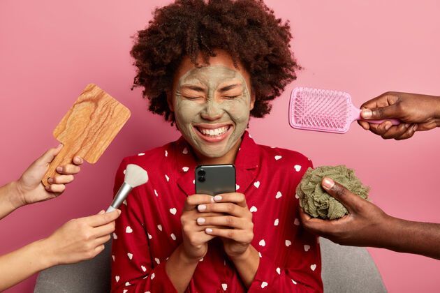化妆快乐的美国黑人妇女使用手机 在线阅读美容博客 笑容灿烂 涂上天然粘土面膜 穿着睡衣坐着请美容师